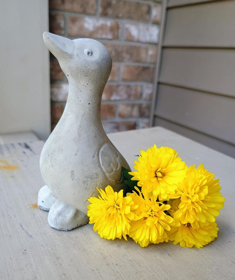 May include: A concrete duck ornament with a weathered, textured finish. The duck is light grey and has a long neck and a slightly open beak. Bright yellow flowers are arranged beside the duck, providing a vibrant contrast.