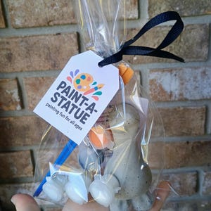 May include: A clear cellophane bag tied with a black ribbon, containing a paint-a-statue kit. The kit includes a small, unpainted statue, paint pots, and a paintbrush. A white tag with the words "PAINT-A-STATUE" is attached.