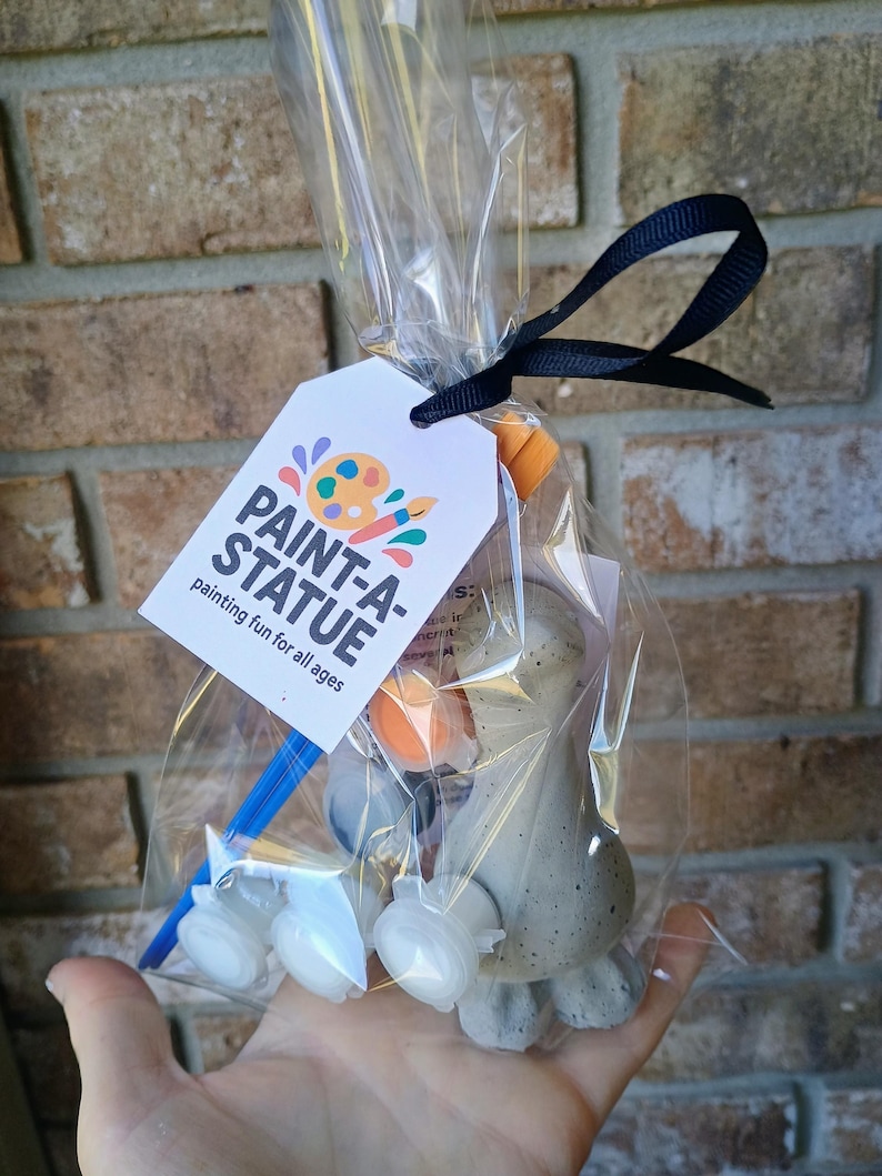 May include: A clear cellophane bag tied with a black ribbon, containing a paint-a-statue kit. The kit includes a small, unpainted statue, paint pots, and a paintbrush. A white tag with the words "PAINT-A-STATUE" is attached.