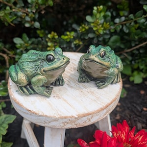 May include: Two green frog figurines with textured skin and dark eyes, positioned on a small, round, white-washed wooden stool. The frogs have a mottled green and gold finish, with light-coloured underbellies and detailed webbed feet.