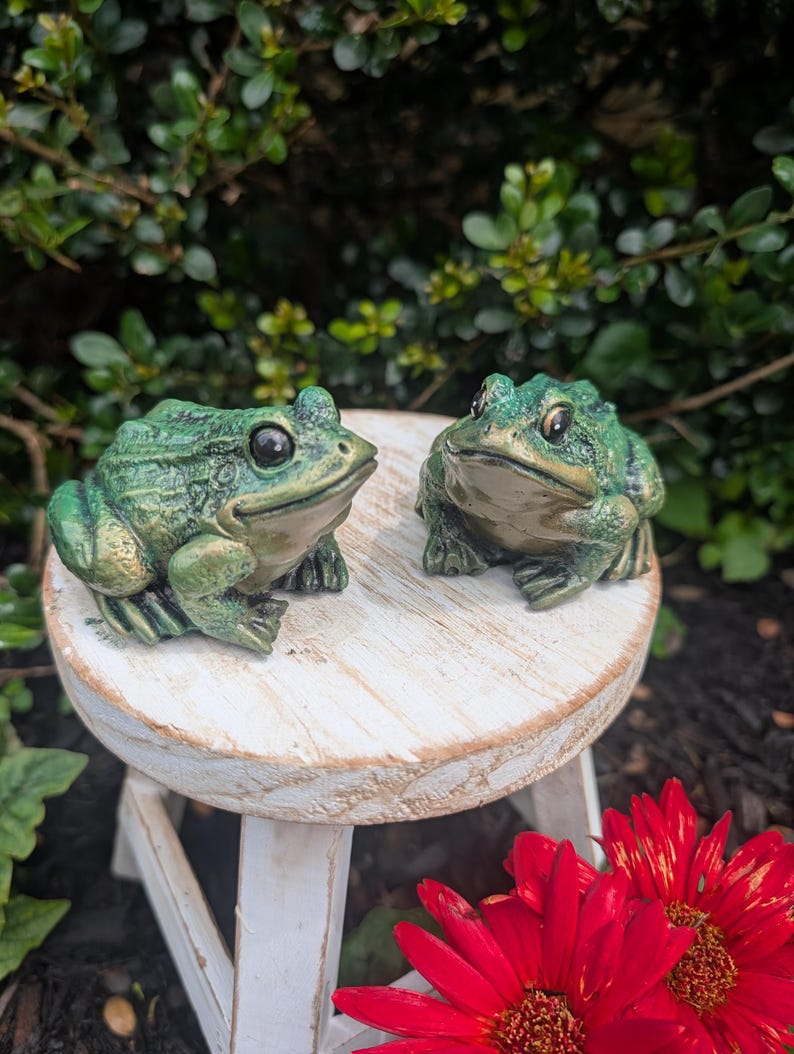 May include: Two green frog figurines with textured skin and dark eyes, positioned on a small, round, white-washed wooden stool. The frogs have a mottled green and gold finish, with light-coloured underbellies and detailed webbed feet.