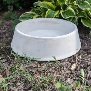 May include: A white ceramic pet bowl with a raised rim sits on a bed of brown mulch and green grass.