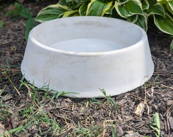 May include: A white ceramic pet bowl with a raised rim sits on a bed of brown mulch and green grass.