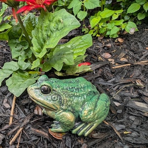 May include: A green frog figurine with a textured, bumpy surface and dark, prominent eyes. The frog is depicted in a seated, resting position with its front legs tucked and back legs folded, showcasing a metallic sheen on its skin.