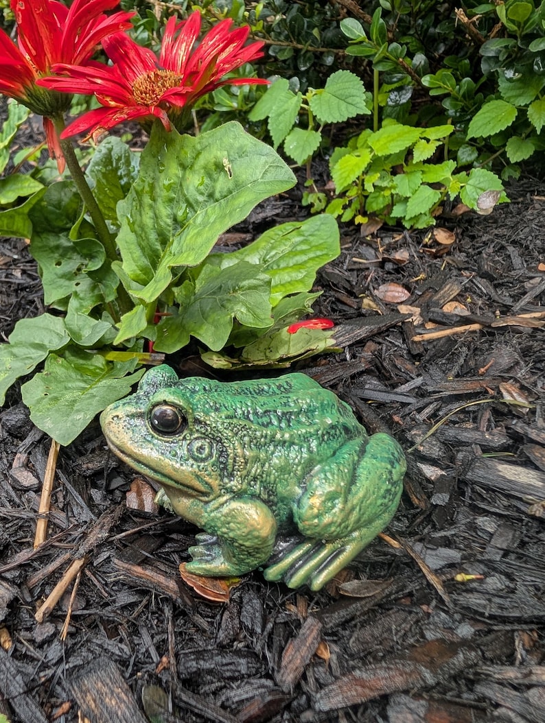 May include: A green frog figurine with a textured, bumpy surface and dark, prominent eyes. The frog is depicted in a seated, resting position with its front legs tucked and back legs folded, showcasing a metallic sheen on its skin.