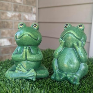 May include: Two green ceramic frogs sitting on a bed of green grass. One frog is in a yoga pose with its hands clasped together. The other frog is sitting with its hands on its chin.