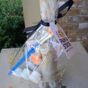 May include: A clear cellophane bag tied with a black ribbon contains a paint-a-statue kit. The kit includes a small statue, paint pots, and a paintbrush. The tag reads "PAINT A STATUE" with a colorful logo. The statue is a light grey colour.