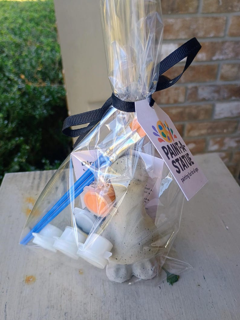 May include: A clear cellophane bag tied with a black ribbon contains a paint-a-statue kit. The kit includes a small statue, paint pots, and a paintbrush. The tag reads "PAINT A STATUE" with a colorful logo. The statue is a light grey colour.