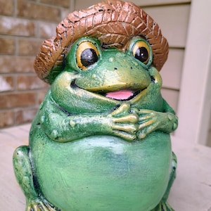 May include: A whimsical green frog statue with a straw hat. The frog has large, expressive eyes, a smiling mouth, and folded hands. The statue is painted with shades of green, gold, and brown, and is a charming garden decoration.