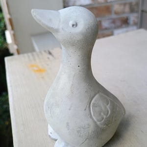 May include: A light grey concrete duck ornament. The duck has a detailed beak, eye, and wing design. This decorative statue is suitable for home or garden use.