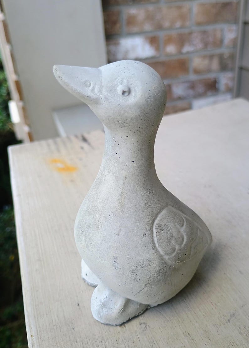 May include: A light grey concrete duck ornament. The duck has a detailed beak, eye, and wing design. This decorative statue is suitable for home or garden use.