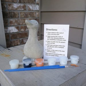 May include: A concrete goose statue with a paint kit. The kit includes small paint pots in various colors, a paintbrush, and instructions. The statue is gray and ready to be painted. The instructions are printed on a white card.