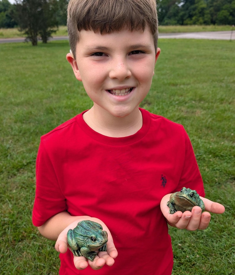 May include: Two small, realistic green frog figurines with textured skin and dark markings. Each frog is held in a palm, showcasing detailed eyes and a crouched posture. The figurines are crafted with a matte finish and deep green tones, capturing the appearance of amphibians in a stationary, lifelike pose.