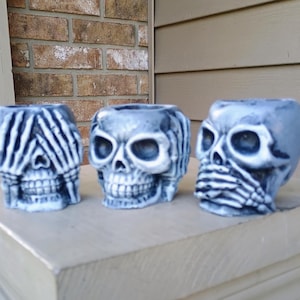 May include: Three gray skull-shaped planters. Each skull has a different hand gesture: covering the eyes, covering the mouth, and covering the mouth. The planters are made of a textured material and are displayed on a light-colored surface.