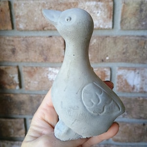 May include: A small, grey concrete duck statue. The duck has a smooth body with a detailed wing design and a long neck. The statue is held in a hand, and the background is a brick wall.