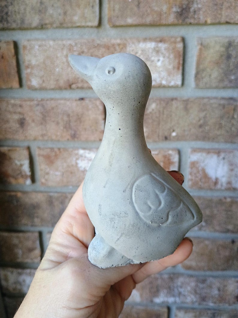 May include: A small, grey concrete duck statue. The duck has a smooth body with a detailed wing design and a long neck. The statue is held in a hand, and the background is a brick wall.