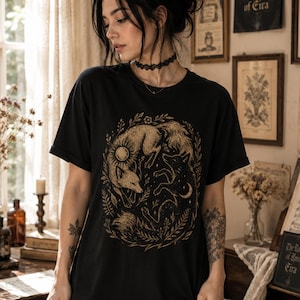 Puede incluir: Una camiseta negra de manga corta con un estampado gráfico en tono dorado de dos zorros entrelazados dentro de una corona floral circular. Un zorro tiene un motivo de sol, mientras que el otro presenta un diseño de luna creciente y estrella.