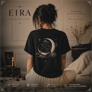 Puede incluir: Una camiseta negra con un estampado gráfico en la espalda de un gato sentado sobre una luna creciente rodeado de nubes y estrellas. El texto EIRA WEAR THE STORY está impreso en la parte delantera, junto a la frase Some stories are written in silence.