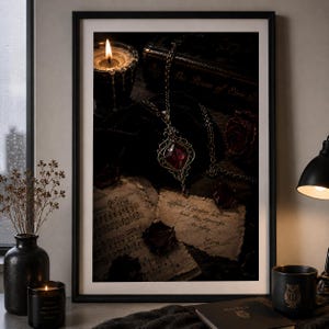 Puede incluir: Una lámina de arte mural enmarcada con una composición oscura y atmosférica que muestra un collar con colgante plateado y una piedra preciosa roja, sobre pergaminos envejecidos con texto escrito a mano, un libro antiguo y pétalos de rosa.