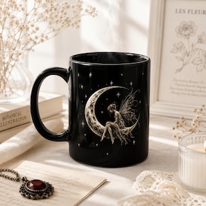 Puede incluir: Una taza de cerámica negra con una ilustración detallada de un hada alada sentada sobre una luna creciente rodeada de pequeñas estrellas. El diseño está representado en un estilo grabado claro sobre la superficie oscura de la taza con asa.