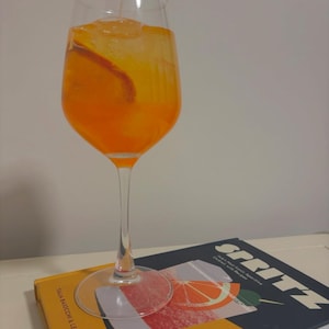 May include: A glass of orange-coloured cocktail with ice and an orange slice in a clear wine glass. The glass is sitting on a book with the word "SPRITZ" printed on the cover.