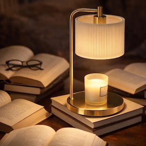 May include: A gold-toned lamp with a cylindrical, ribbed shade illuminates a lit candle on a stack of books. The lamp has a curved arm and a circular base. Several open books and a pair of glasses are also visible.