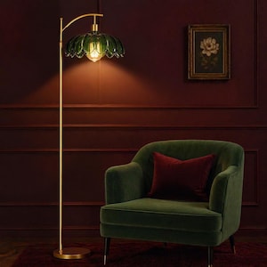 May include: A gold floor lamp with a green glass shade shaped like a flower. The lamp is next to a green velvet armchair with a burgundy pillow. A framed floral print hangs on the burgundy wall.