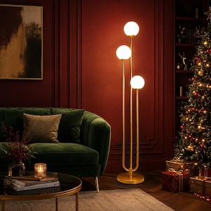 May include: A floor lamp featuring a gold-toned metal stand with three vertical poles of varying heights, each topped with a spherical white glass globe light. The lamp has a circular base and emits a warm glow from the three illuminated bulbs.