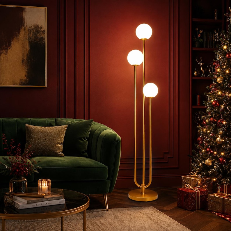 May include: A floor lamp featuring a gold-toned metal stand with three vertical poles of varying heights, each topped with a spherical white glass globe light. The lamp has a circular base and emits a warm glow from the three illuminated bulbs.