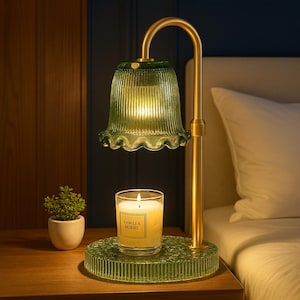 May include: A green glass lamp with a gold-colored metal frame. The lamp has a scalloped shade and a round base. A lit candle in a glass jar sits on the base. A small potted plant is to the left.