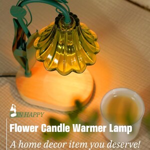 May include: A flower-shaped candle warmer lamp with a green metal stem and leaves. The lamp has a yellow glass shade and sits on a wooden base. A white ceramic cup is next to the lamp.