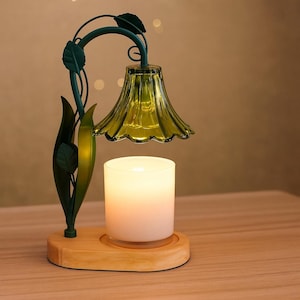 May include: A decorative candle warmer with a green glass shade shaped like a flower. The metal frame is green with leaf accents. The candle sits on a wooden base, and the candle is white.