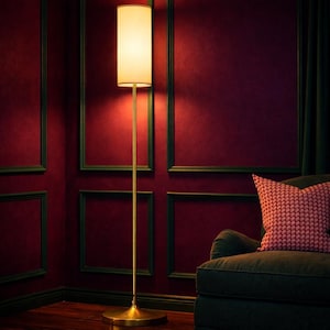 May include: A tall, gold-toned floor lamp with a cream-colored shade illuminates a room with burgundy walls and dark green trim. A patterned red pillow rests on a dark green armchair in the corner. The lamp's base is circular.