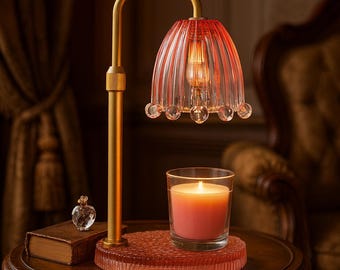 May include: A decorative table lamp with a red glass shade and a gold-colored metal frame. A lit candle in a glass jar sits on a matching red base. A small perfume bottle and a book are also on the table.