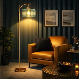 May include: A gold floor lamp with a teal glass shade illuminates a room. The lamp has a curved arm and a round base. The room features a brown leather armchair, a black pillow, and a small side table. Two framed pictures hang on the wall.