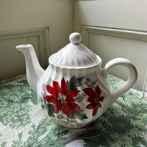 May include: White ceramic teapot with a lid and handle. The teapot is decorated with red poinsettia flowers and green leaves. The teapot is on a green and white patterned tablecloth.