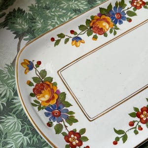 May include: A rectangular white serving tray with a floral design. The tray features a gold trim and is decorated with colorful flowers in shades of red, yellow, and blue. The flowers are arranged along the edges.