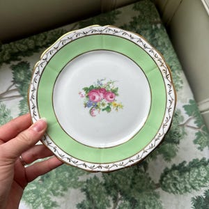 Puede incluir: Un plato pequeño y decorativo con un borde verde claro y un diseño floral en el centro. El plato tiene un borde festoneado con ribete dorado. El centro presenta rosas rosas y otras flores coloridas. El plato es sostenido por una persona.
