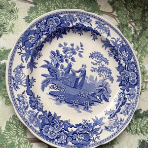 Op de afbeelding: Een wit en blauw keramisch bord met een bloemen- en pastorale ontwerp. Het bord heeft een blauwe bloemenrand en een centrale scène van figuren bij een put. Het bord heeft een geschulpte rand.