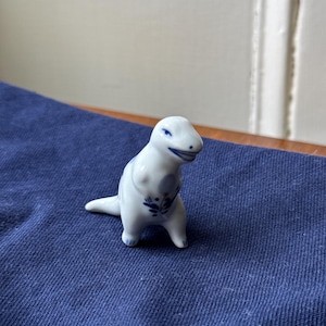 May include: A small white ceramic dinosaur figurine with blue and white painted details. The dinosaur is standing on a blue surface.