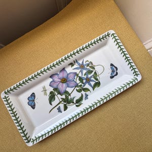 May include: White rectangular ceramic serving tray with a botanical design. The tray has a green leaf border and a central illustration of purple and blue flowers, butterflies, and a dragonfly.