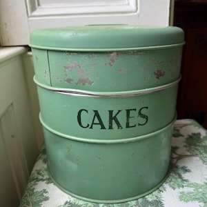 Peut inclure: Une boîte à gâteaux vintage en métal vert à trois niveaux avec le mot "CAKES" en lettres noires. La boîte montre des signes d'usure, avec quelques pertes de peinture. Le récipient à gâteaux est rond et possède un couvercle.