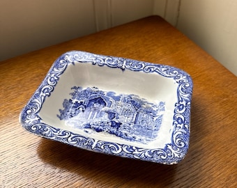 May include: A small, white ceramic dish with a blue and white floral pattern. The dish has a rectangular shape and a slightly raised rim.