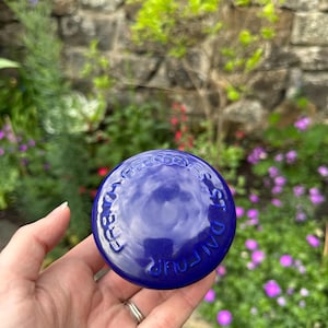 May include: A deep blue, round ceramic lid with embossed text. The lid is held in a hand, with a garden background featuring green foliage and purple flowers. The text on the lid reads "CREAM FOUR STOKE ON TRENT".