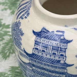 May include: A white ceramic jar with a blue Willow pattern design. The jar features a detailed illustration of a pagoda, trees, and other elements of the classic blue and white pattern. The jar has a rounded shape and a wide opening.