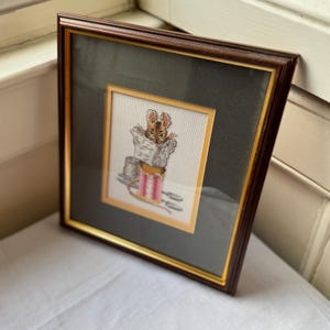 Könnte beinhalten: Gerahmtes Kreuzstich-Kunstwerk mit einer braunen Maus, die eine Zeitung liest. Das Kunstwerk ist in einem goldfarbenen Rahmen mit einem dunkelgrünen Passepartout und einem dunkelbraunen Holzrahmen eingefasst. Die Maus ist in Braun-, Grau- und Rosatönen dargestellt.