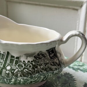 May include: Cream gravy boat with a green transferware design. The illustration shows a horse-drawn carriage and figures, with a sign reading "Swan". The handle is curved and the interior is plain.