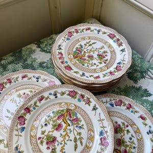 May include: A stack of round ceramic plates featuring a cream-colored center with a central tree motif in shades of pink, green, and blue. The rim is decorated with a repeating floral pattern of pink roses and blue berries on a white background.