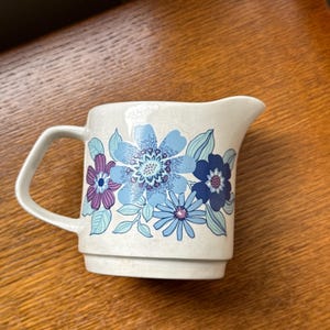 Könnte beinhalten: Cremefarbener Keramik-Milchkännchen mit Henkel und Ausguss. Verziert mit blauen, lila und türkisfarbenen Blumenmustern. Die Blumen sind in verschiedenen Größen und Stilen gehalten, was dem Design einen Vintage-Touch verleiht. Auf einer Holzoberfläche.