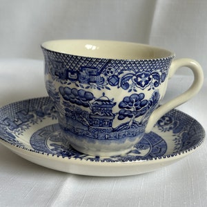 May include: A blue and white teacup and saucer set featuring a traditional Chinese design. The cup and saucer are both decorated with a blue and white pattern of a pagoda, trees, and flowers.